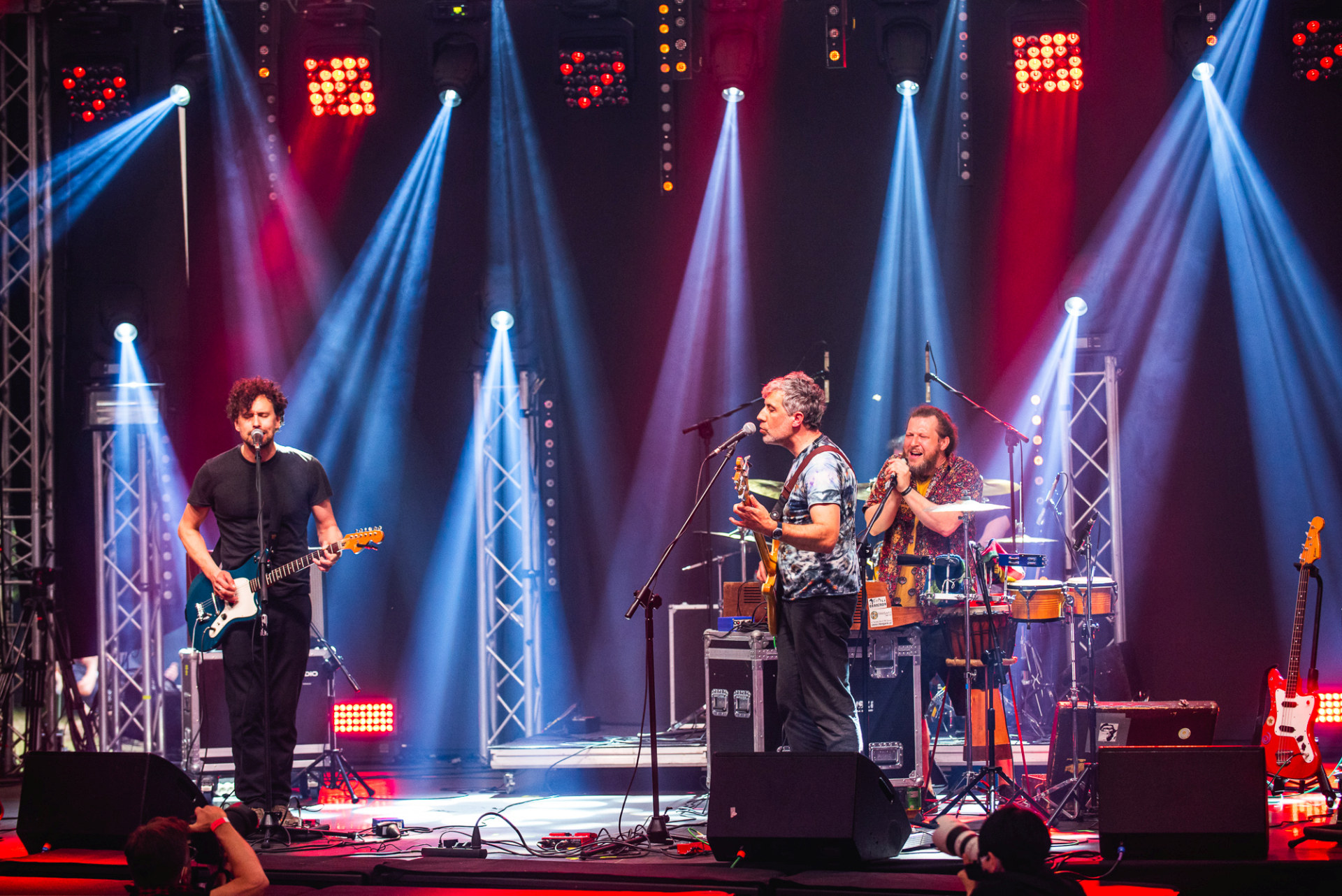 Na scenie są kolejno gitarzysta, wokalista i perkusista. Wszyscy mężczyźni śpiewają.