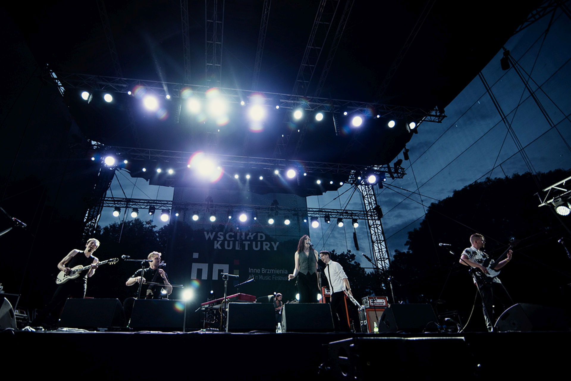 Na środku zdjęcia wokalistka. W tle są gitarzyści, perkusista i wiolonczelista.