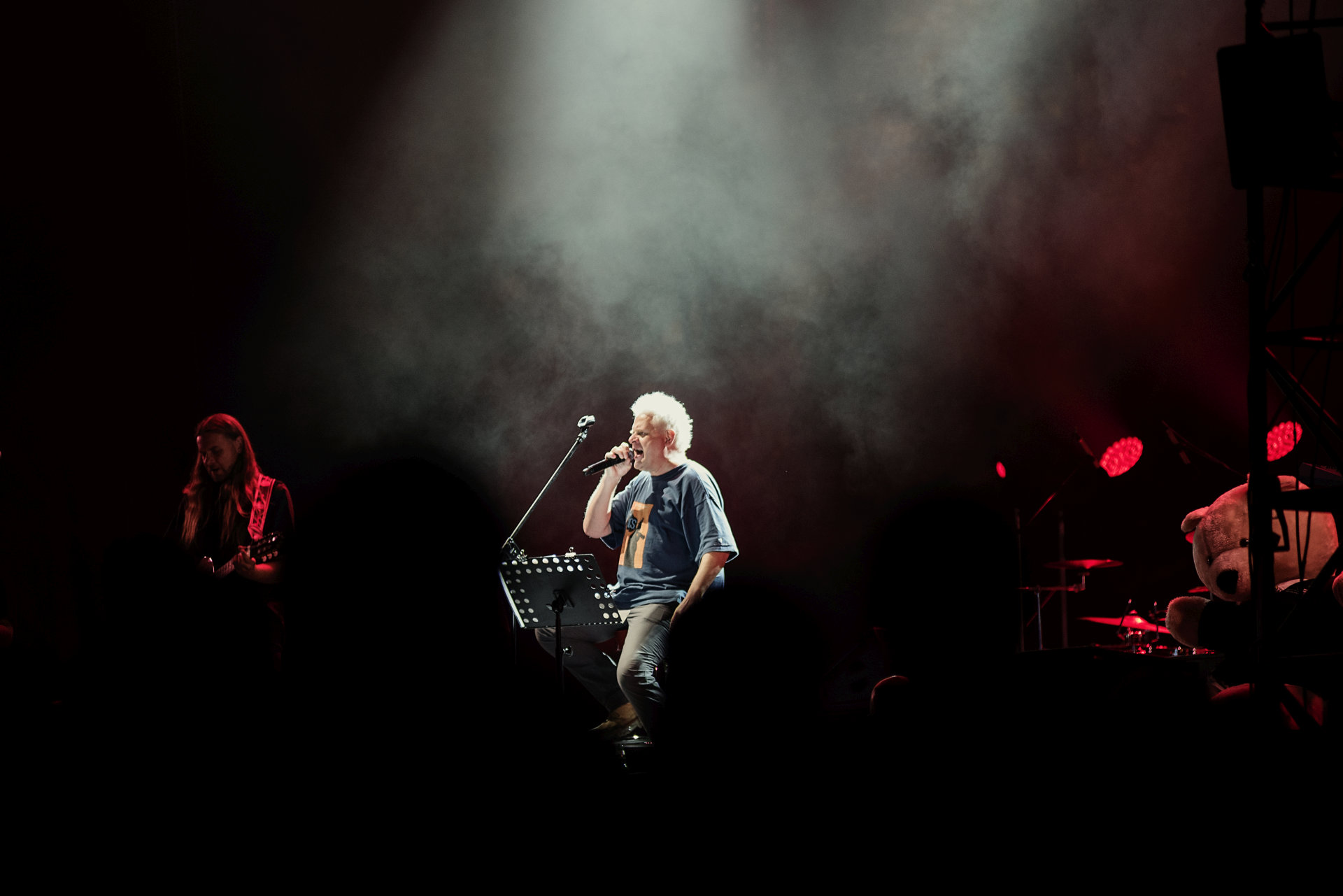 Na scenie jest wokalista i gitarzysta. Przy perkusji siedzi pluszowy miś.