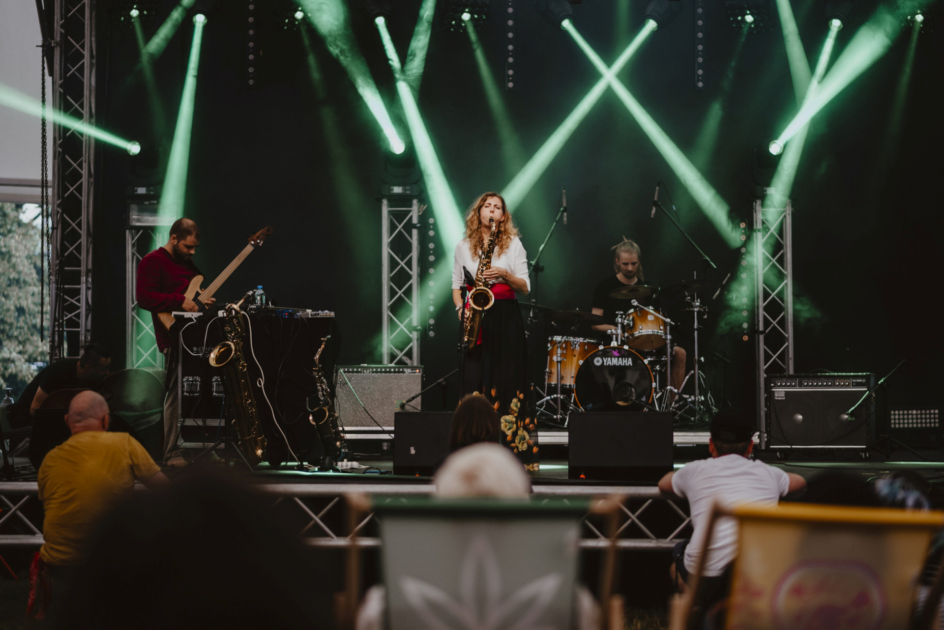 Na scenie kolejno gitarzysta, saksofonistka i perkusista. Pod sceną na leżakach siedzi publiczność.